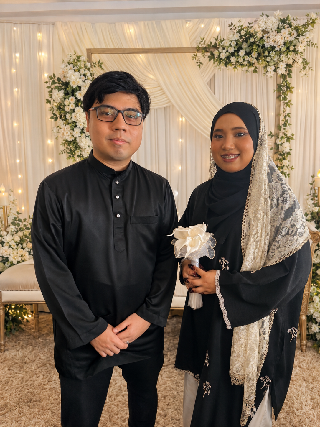 Athirah and Syamir dressed in black before an elegant white floral backdrop